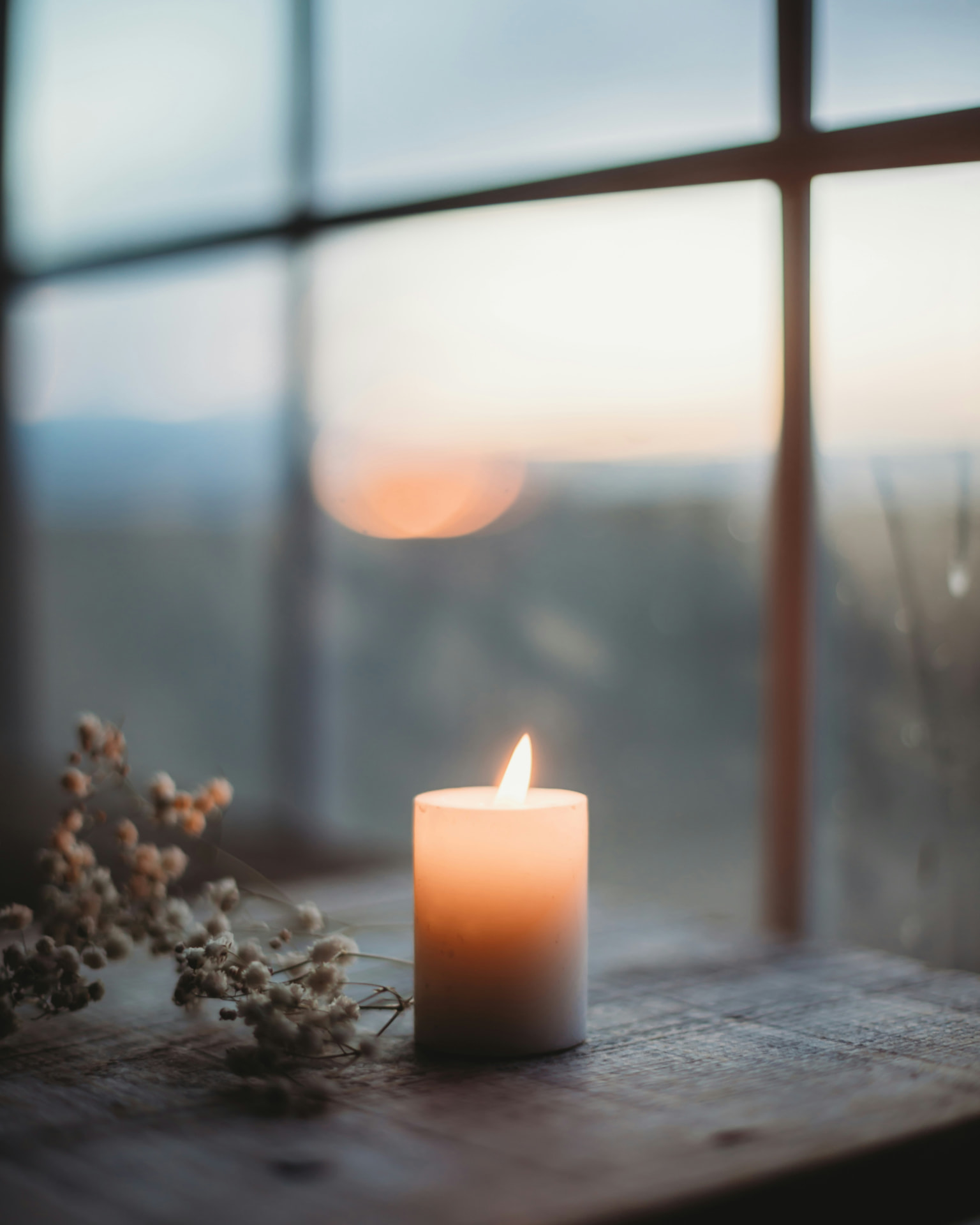 A candle burning quietly on a wooden table beside a window at dusk